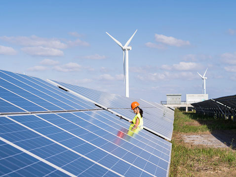 Een vrouw met bouwhelm loopt langs een veld met zonnepanelen met windmolens op de achtergrond